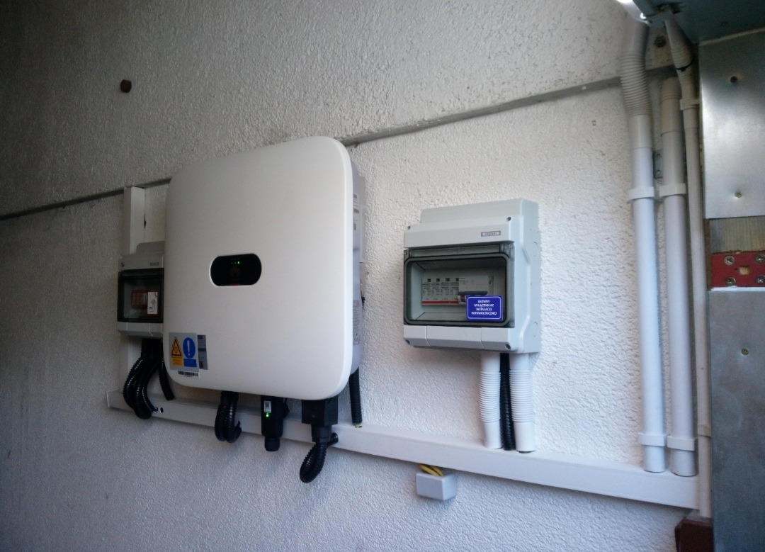 Biały inwerter fotowoltaiczny i skrzynka elektryczna zamontowane na ścianie o szorstkiej fakturze. Widoczne kable i rury instalacyjne.