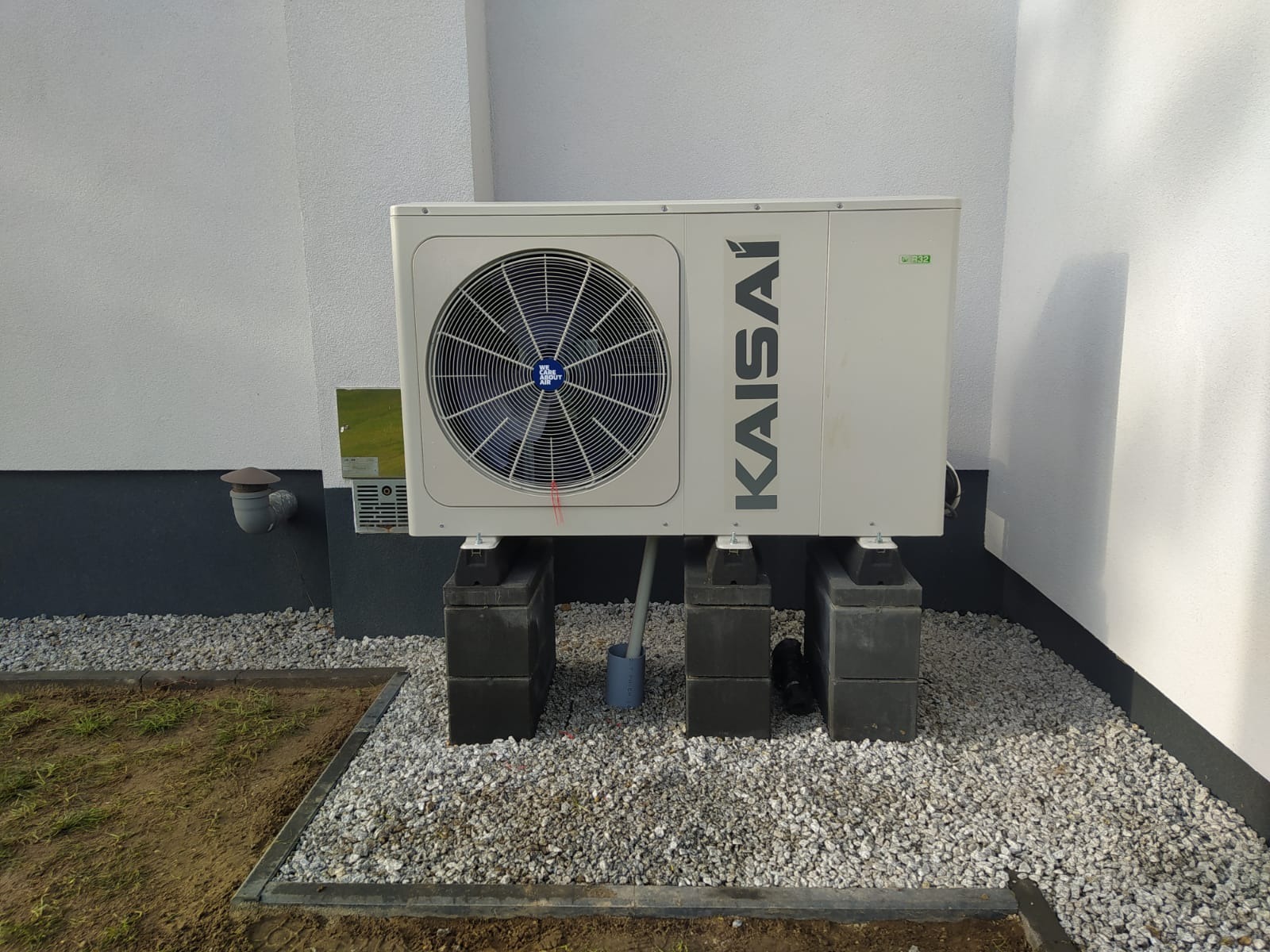 Biała pompa ciepła Kaisai zamontowana na betonowych blokach przy ścianie budynku, otoczona drobnym żwirem i obrzeżem trawnika. Widoczne elementy instalacji.