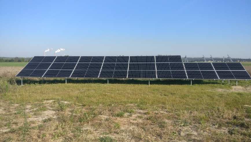 Rząd paneli słonecznych na metalowych wspornikach, na tle błękitnego nieba i odległych kominów. Instalacja fotowoltaiczna na łące, widok z poziomu gruntu.
