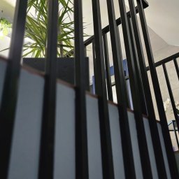 FUZJA DEVELOPMENT MIKOŁAJ SIKORSKI - Nowoczesna balustrada schodowa z czarnych, pionowych elementów, widziana z dołu, z rośliną w tle. Minimalistyczny design i neutralna kolorystyka.