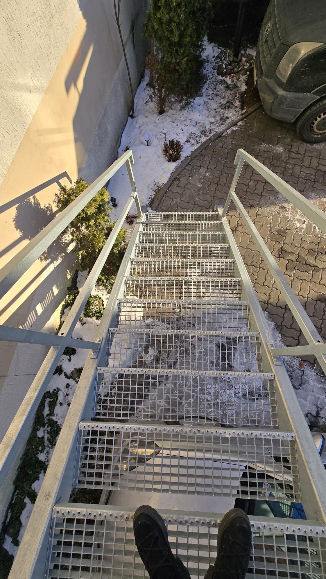 Zewnętrzne schody metalowe z ażurowymi stopniami i poręczami, prowadzące w górę. Widok z góry na stopnie, z butami osoby fotografującej w dolnej części kadru.