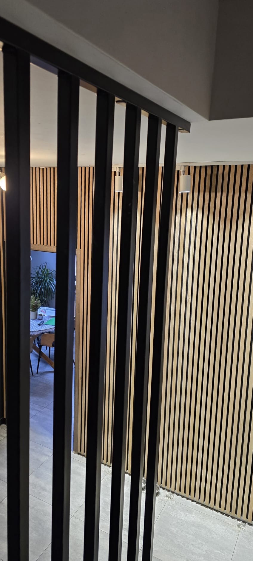 Nowoczesna, czarna ścianka działowa z pionowych profili na tle drewnianej okładziny ściennej. Widok na wnętrze przez szczeliny.