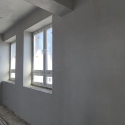 Perfekt Tynk - Wnętrze po tynkowaniu maszynowym: gładkie, szare ściany z dwoma oknami w białych ramach. Widoczne kable na podłodze, surowy stan wykończenia.