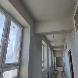 Perfekt Tynk - Korytarz po tynkowaniu maszynowym: gładkie, szare ściany, białe okna z folią ochronną, widoczne instalacje elektryczne na podłodze. Perspektywa w głąb pomieszczenia.