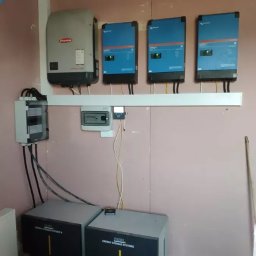 Instalacja wewnętrzna: dwa akumulatory energii, falownik Fronius i trzy inwertery Victron na ścianie z różową płytą gipsową. Widoczne kable i skrzynka elektryczna.