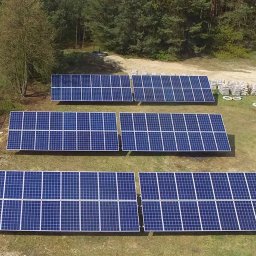 Zestaw paneli słonecznych na trawiastym terenie, widok z góry. Panele ułożone w rzędach, w tle las i worki z materiałem budowlanym.