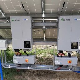 Dwa szare inwertery Growatt zamontowane pod panelami słonecznymi, z widocznymi przewodami i skrzynkami przyłączeniowymi. Instalacja na zewnątrz, w tle trawa.