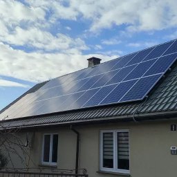 Panele na dachu domu jednorodzinnego z szarej dachówki, błękitne niebo z chmurami w tle. Widoczne okno i numer domu.