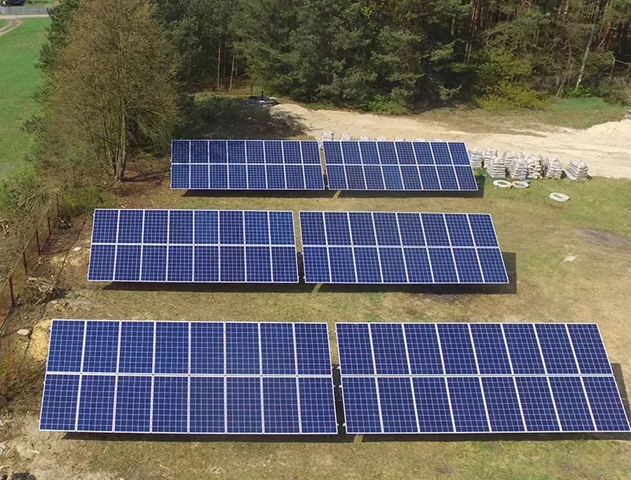 Zestaw paneli słonecznych na trawiastym terenie, widok z góry. Panele ułożone w rzędach, w tle las i worki z materiałem budowlanym.