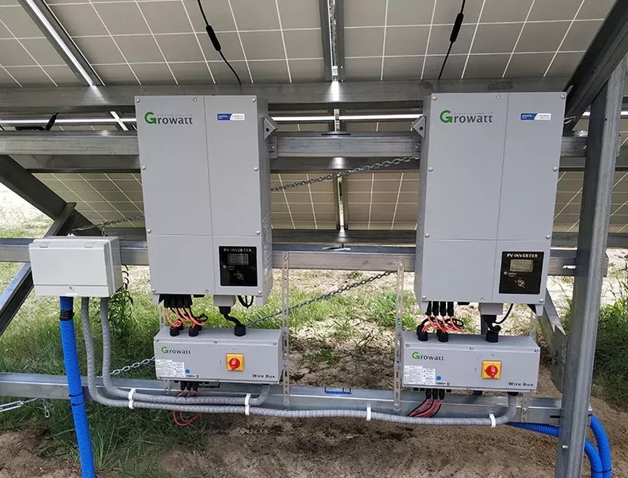 Dwa szare inwertery Growatt zamontowane pod panelami słonecznymi, z widocznymi przewodami i skrzynkami przyłączeniowymi. Instalacja na zewnątrz, w tle trawa.