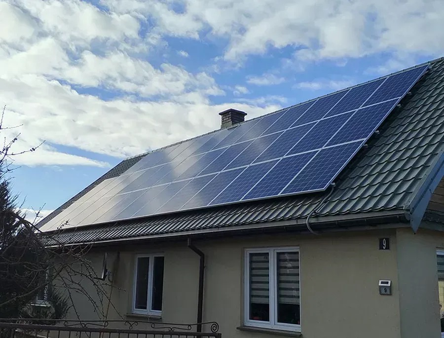 Panele na dachu domu jednorodzinnego z szarej dachówki, błękitne niebo z chmurami w tle. Widoczne okno i numer domu.