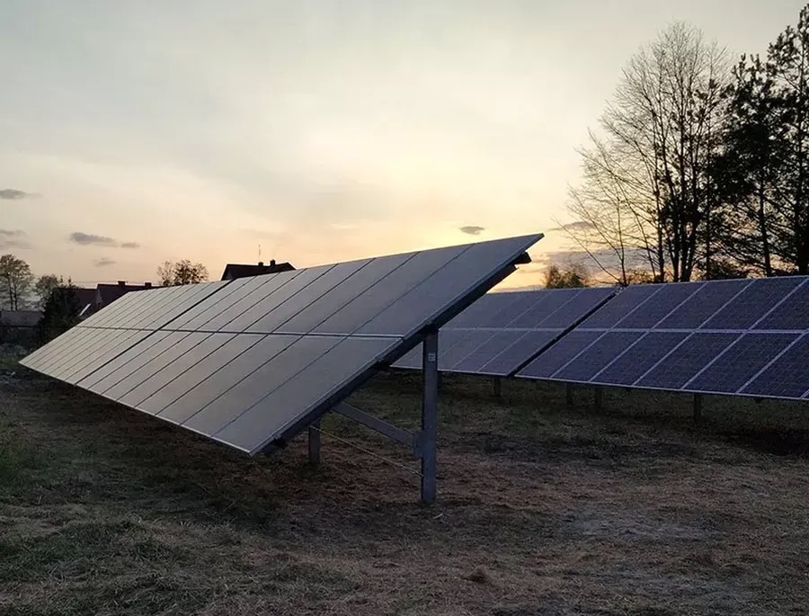Instalacja paneli słonecznych na gruncie o zmierzchu. Widoczne rzędy paneli, konstrukcja wsporcza i budynki w tle. Delikatne światło zachodzącego słońca.