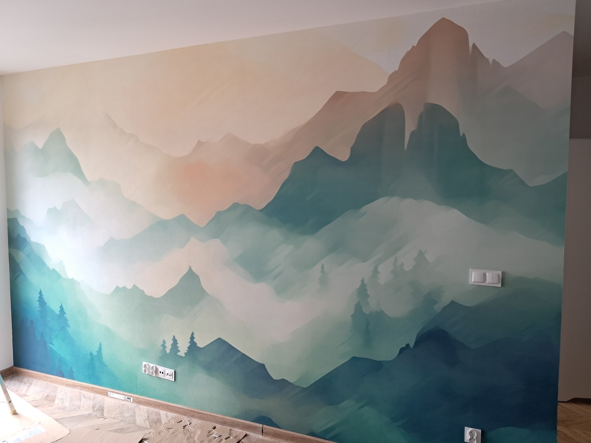 Ściana z malowidłem górskim w odcieniach zieleni i beżu, widoczne gniazdka elektryczne i fragment podłogi z zabezpieczeniami, sugerujące świeżo zakończone prace.