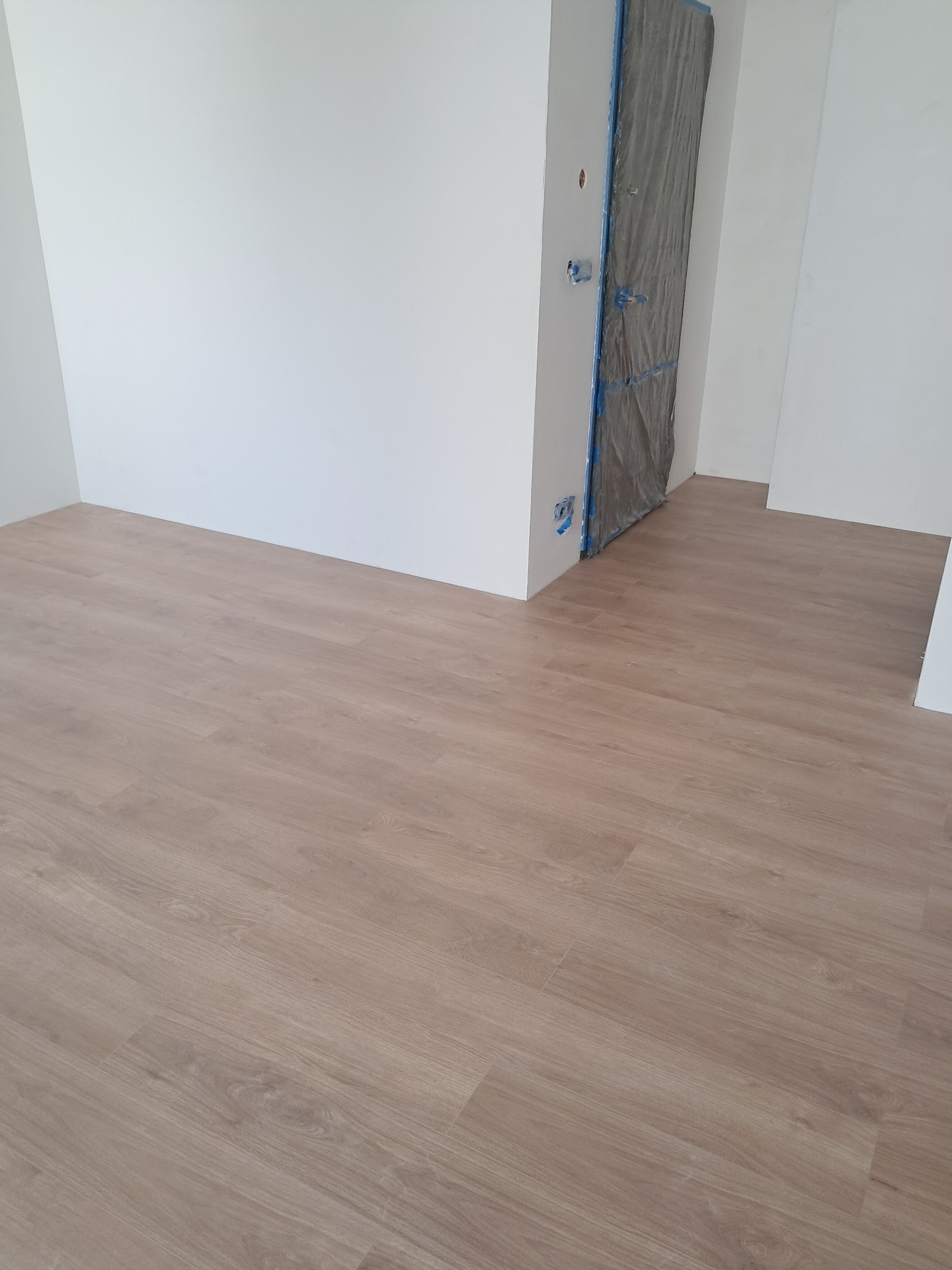 Jasne panele podłogowe w nowym wnętrzu, ściany pomalowane na biało, zabezpieczone drzwi folią malarską. Widok na świeżo wykończone pomieszczenie.