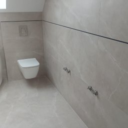 Wykończona łazienka na poddaszu z beżowymi płytkami imitującymi marmur, wiszącą toaletą i chromowanymi zaworami. Minimalistyczny design z czarnymi akcentami.