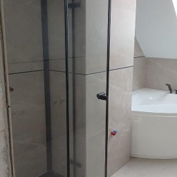 Nowoczesna kabina prysznicowa z czarnymi profilami i szklanymi ścianami, w łazience z beżowymi płytkami imitującymi marmur i białą wanną narożną.