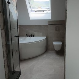 Wykończona łazienka na poddaszu z oknem dachowym. Uwagę przykuwa narożna wanna, szklana kabina prysznicowa i płytki imitujące marmur. Widok z korytarza.