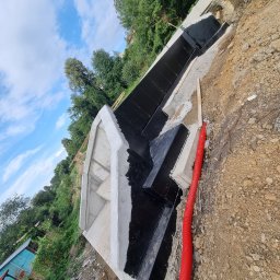 Mateusz Chrobak - Betonowy fundament schodów zabezpieczony czarną izolacją bitumiczną, widoczna czerwona rura karbowana w ziemi, tło z zielenią i błękitnym niebem. Ujęcie z boku.