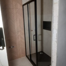 Kabina prysznicowa z czarną ramą w wykończonej łazience. Widoczne płytki w dwóch odcieniach beżu i czarny brodzik. Ściana po prawej w trakcie wykańczania.