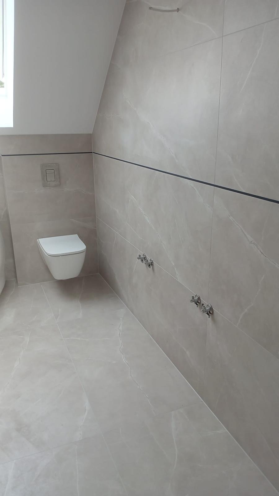 Wykończona łazienka na poddaszu z beżowymi płytkami imitującymi marmur, wiszącą toaletą i chromowanymi zaworami. Minimalistyczny design z czarnymi akcentami.