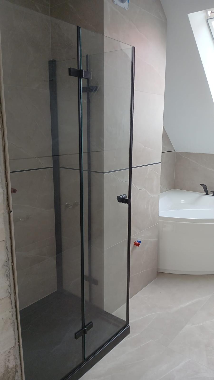 Nowoczesna kabina prysznicowa z czarnymi profilami i szklanymi ścianami, w łazience z beżowymi płytkami imitującymi marmur i białą wanną narożną.