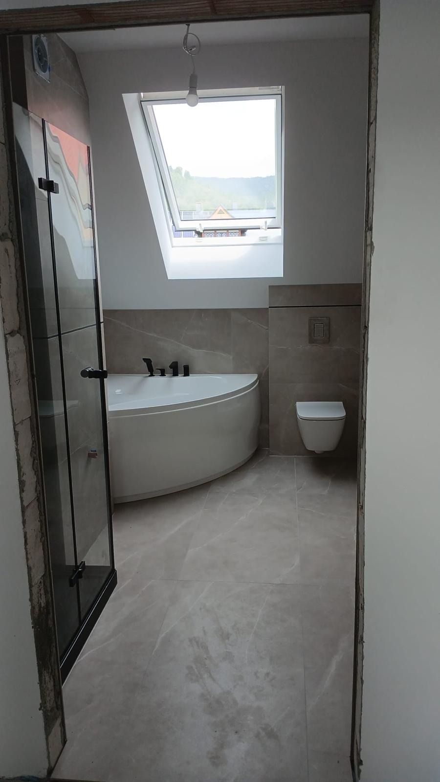 Wykończona łazienka na poddaszu z oknem dachowym. Uwagę przykuwa narożna wanna, szklana kabina prysznicowa i płytki imitujące marmur. Widok z korytarza.