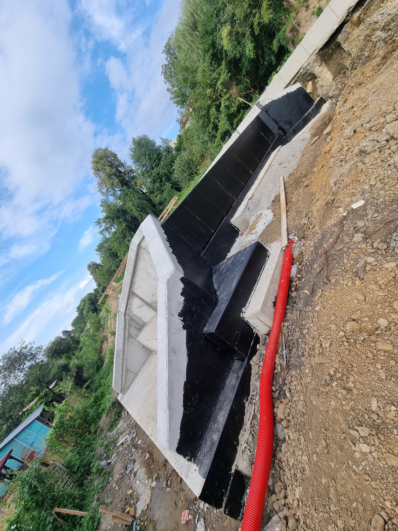 Betonowy fundament schodów zabezpieczony czarną izolacją bitumiczną, widoczna czerwona rura karbowana w ziemi, tło z zielenią i błękitnym niebem. Ujęcie z boku.
