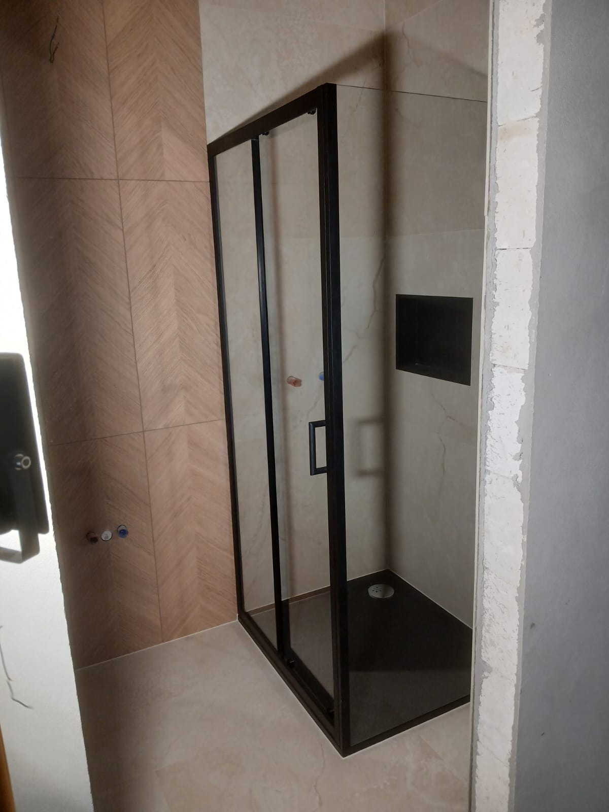 Kabina prysznicowa z czarną ramą w wykończonej łazience. Widoczne płytki w dwóch odcieniach beżu i czarny brodzik. Ściana po prawej w trakcie wykańczania.
