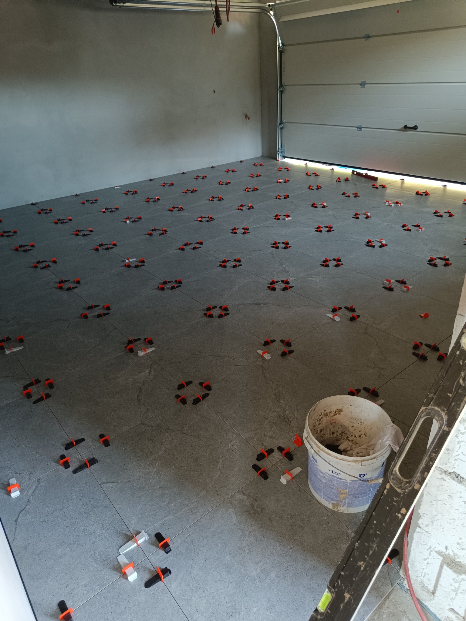Szare płytki podłogowe w garażu, w trakcie układania, z użyciem systemu poziomowania. Widoczny kubeł z zaprawą i poziomica. Oświetlenie garażowe.