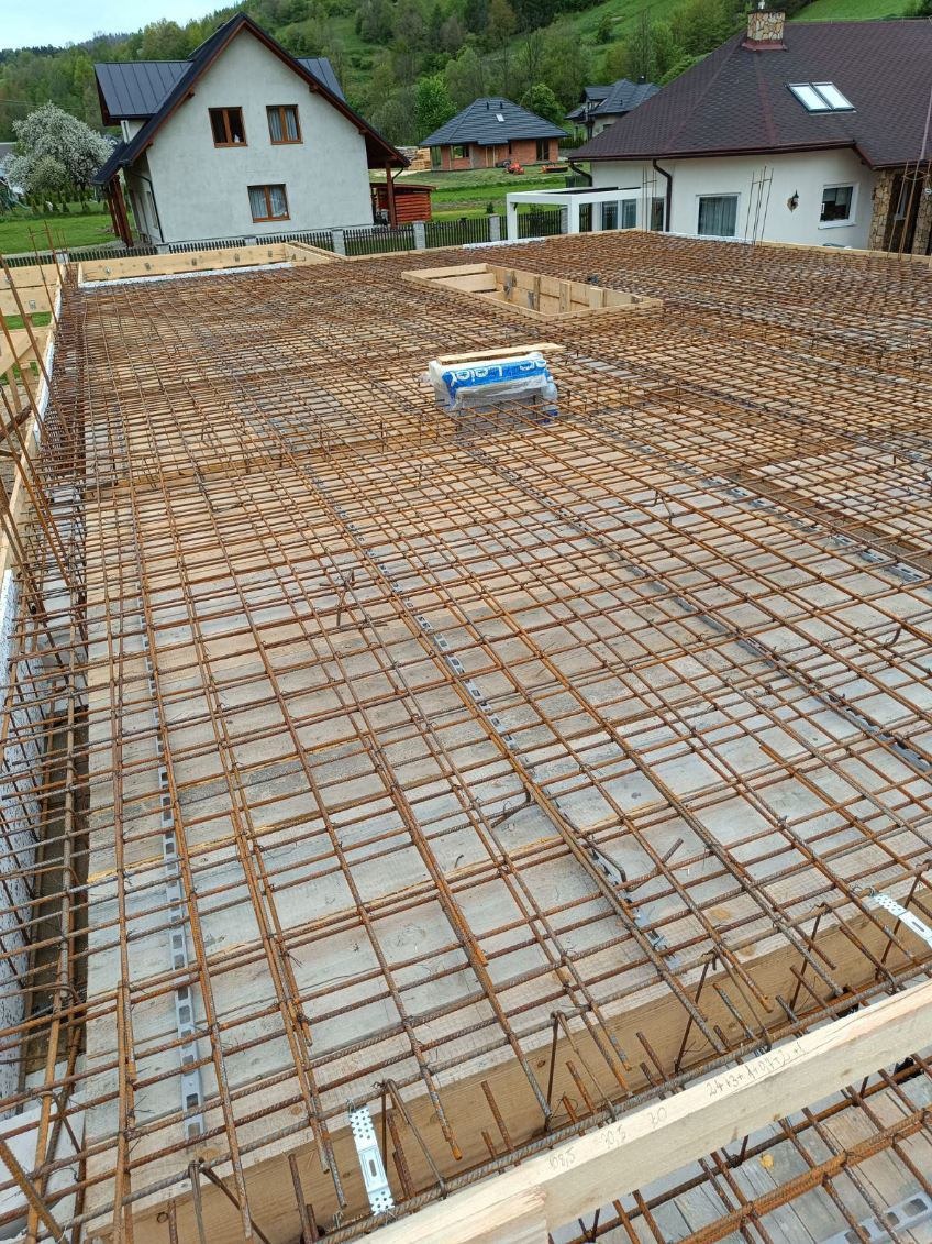 Zbrojenie płyty fundamentowej domu jednorodzinnego z widocznymi elementami szalunku i otaczającym krajobrazem. Siatka z prętów stalowych przygotowana do zalania betonem.