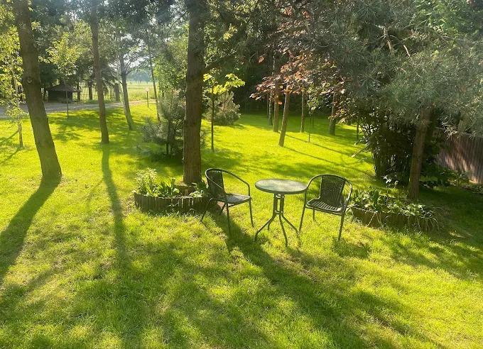 Zadbany trawnik z drzewami i zestawem mebli ogrodowych (stół i dwa krzesła) w słoneczny dzień. Widoczne rabaty kwiatowe i drewniane obrzeża.