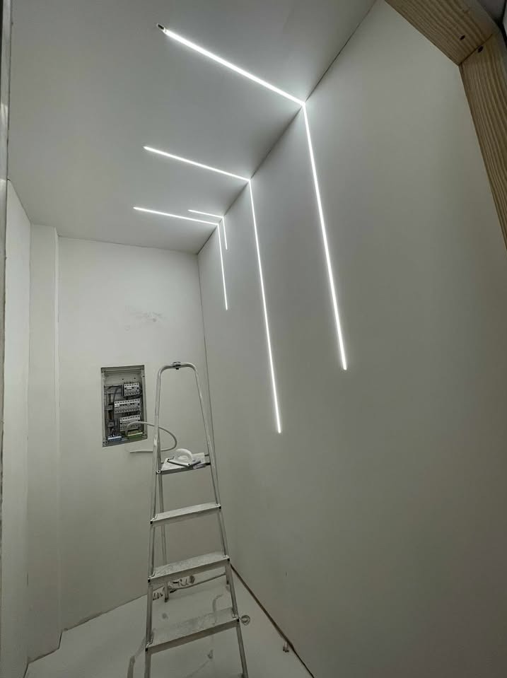 Białe, minimalistyczne wnętrze z liniowym oświetleniem LED w suficie i na ścianach. Widoczna drabina i skrzynka elektryczna w ścianie. Nowoczesny design.