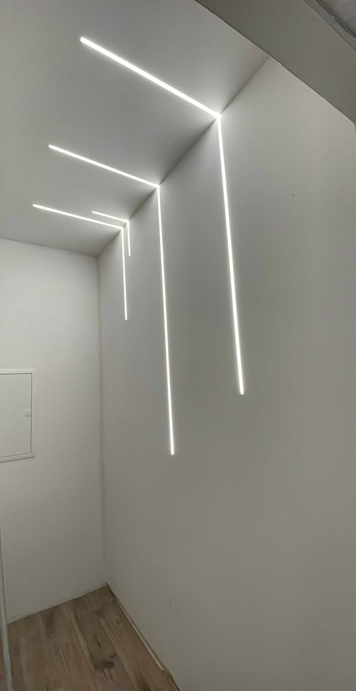 Minimalistyczne oświetlenie LED w rogu pomieszczenia, tworzące geometryczny wzór na białych ścianach i suficie. Widoczny fragment drewnianej podłogi.
