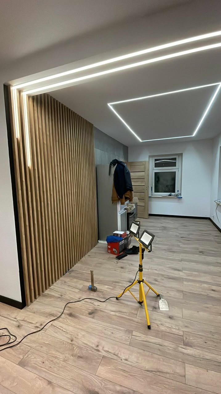 Nowoczesne oświetlenie LED w kształcie kwadratu na suficie i pionowe listwy z podświetleniem LED na ścianie, lampa robocza na trójnogu na podłodze.