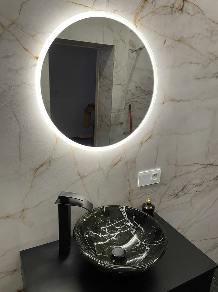 Nowoczesna łazienka z okrągłym lustrem LED, czarną umywalką nablatową i baterią. Marmurowe płytki na ścianie, gniazdko elektryczne. Eleganckie wykończenie wnętrza.