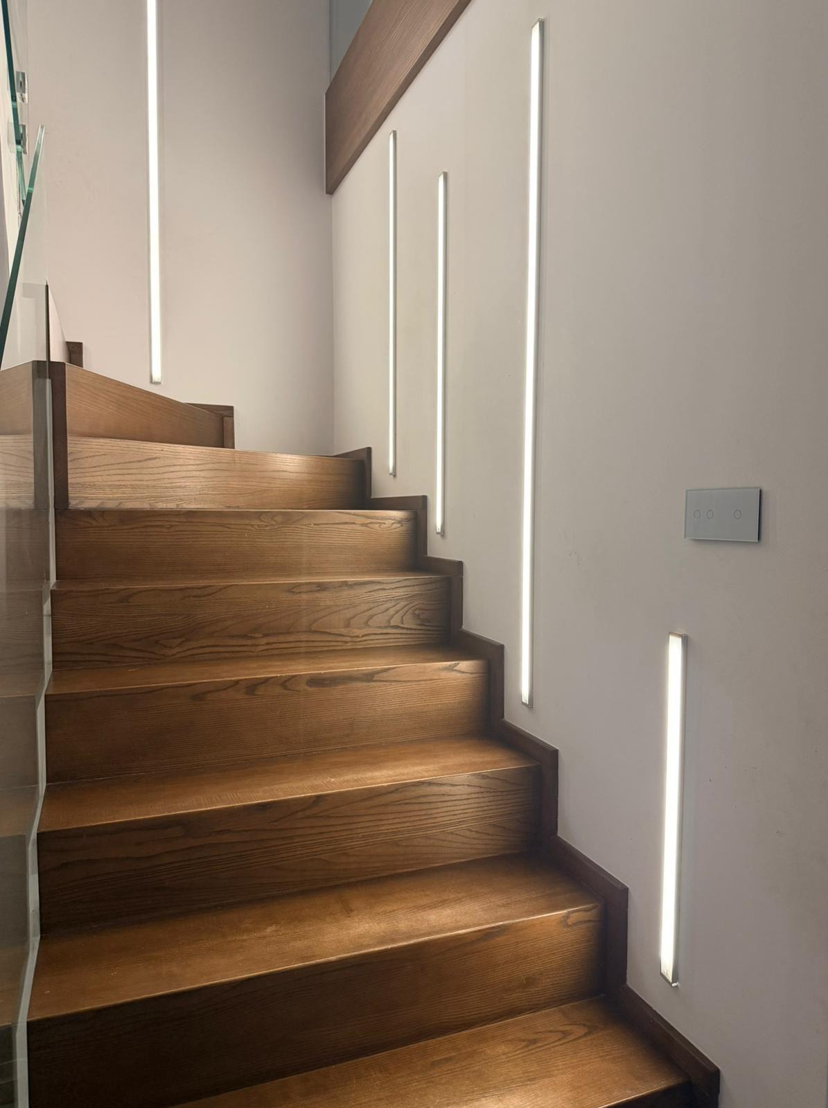 Drewniane schody z podświetleniem LED wzdłuż ściany. Nowoczesne oświetlenie schodowe z włącznikiem dotykowym. Ciepłe światło podkreśla naturalny rysunek drewna.