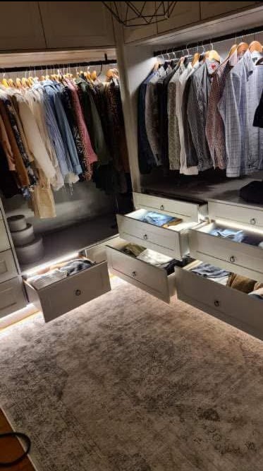 Nowoczesna garderoba z oświetleniem LED w szufladach i pod półkami, ubrania na wieszakach, dywan w odcieniach szarości. Funkcjonalne i stylowe rozwiązanie do przechowywania.