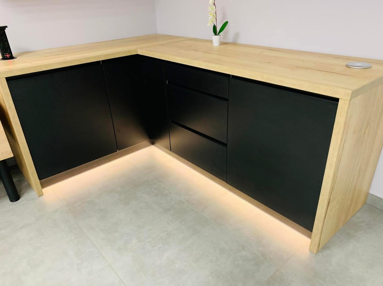 Nowoczesna zabudowa meblowa z czarnymi frontami i drewnianym blatem, podświetlona listwą LED przy podłodze. Minimalistyczny design, jasne płytki podłogowe.