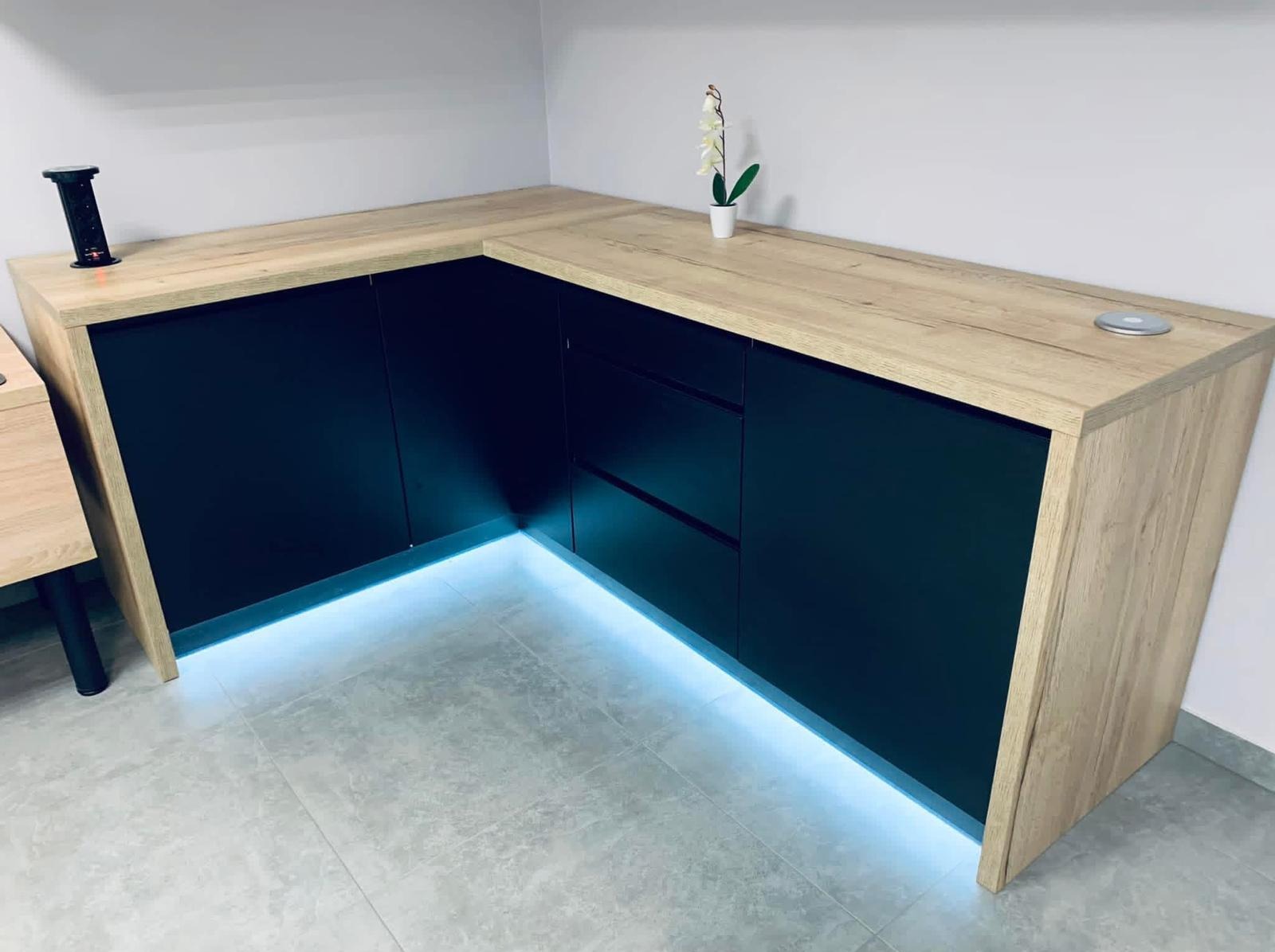 Nowoczesna zabudowa meblowa z czarnymi frontami i blatem w odcieniu jasnego drewna, podświetlona subtelnym, niebieskim światłem LED u dołu szafek.