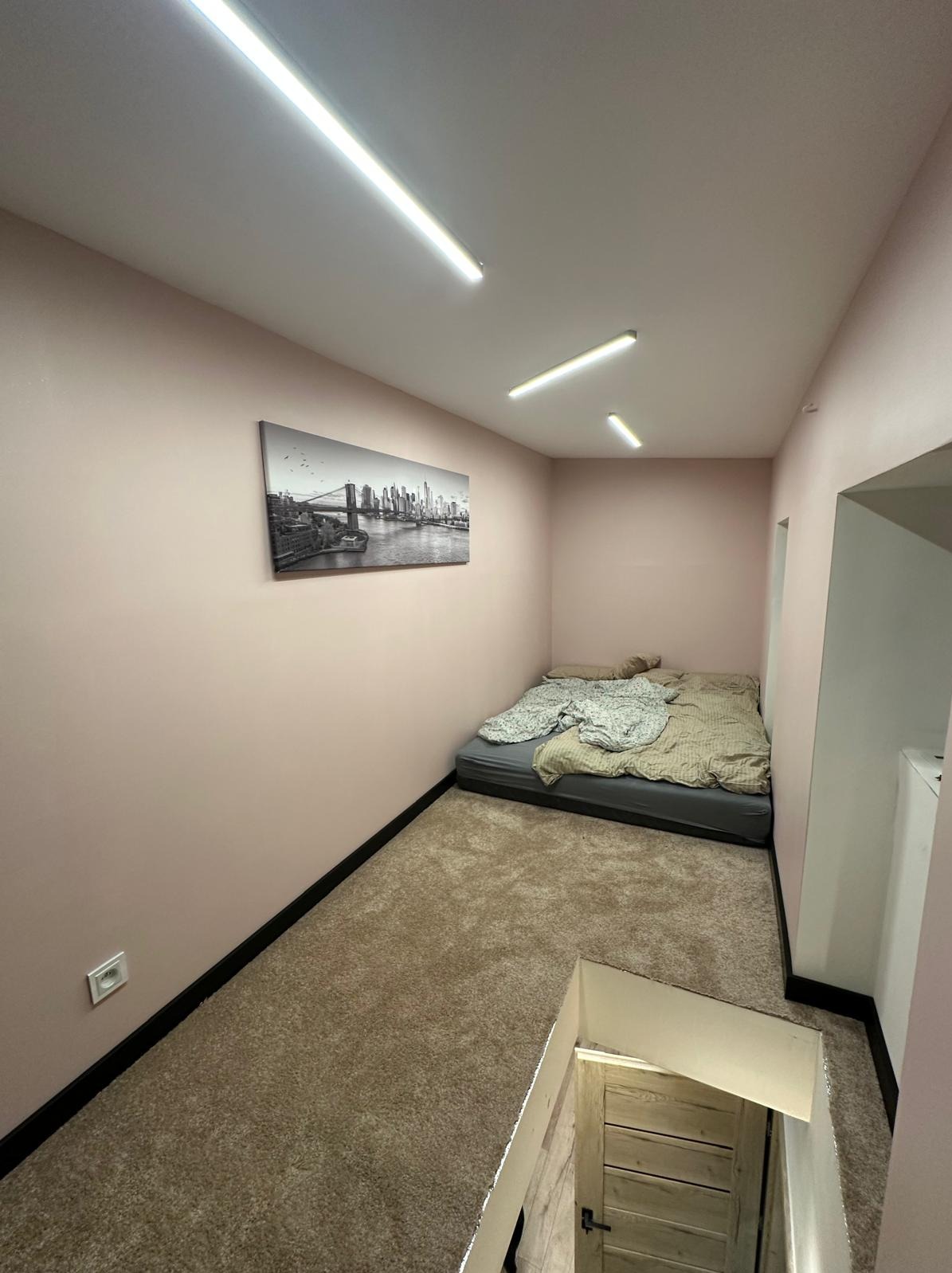 Pokój z łóżkiem na podłodze i obrazem Nowego Jorku na ścianie, oświetlony trzema lampami sufitowymi. Widoczny fragment schodów. Wykończenie wnętrz w odcieniach różu i beżu.