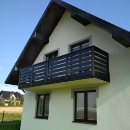 Usługi Stolarskie - Elewacja domu z balkonem z czarną, nowoczesną balustradą. Widok na budynek z zewnątrz w słoneczny dzień. W tle pole i zabudowania.