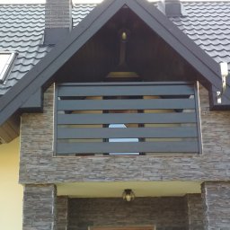 Usługi Stolarskie - Elewacja domu z balkonem z poziomymi, ciemnymi deskami. Widoczny fragment dachu z kominem i drzwi wejściowe. Kamienna okładzina elewacyjna.