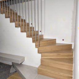 Usługi Stolarskie - Drewniane schody zabiegowe z ażurową balustradą z pionowych, metalowych prętów i drewnianą poręczą, w pomieszczeniu z niewykończonymi ścianami.