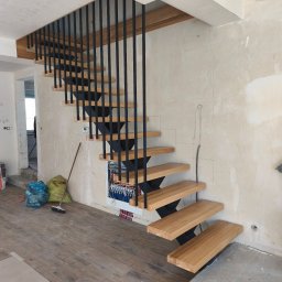 Usługi Stolarskie - Nowoczesne schody dywanowe z drewnianymi stopniami i czarnymi, pionowymi balustradami, zamontowane w pomieszczeniu w trakcie wykończenia, z widoczną instalacją pod spodem.