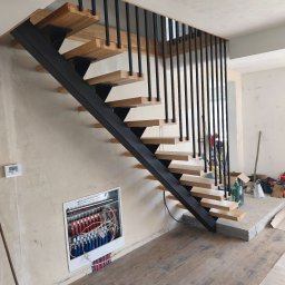 Usługi Stolarskie - Nowoczesne schody z czarną metalową konstrukcją i jasnymi drewnianymi stopniami, z pionowymi czarnymi balustradami. W tle niedokończona ściana z widocznym rozdzielaczem ogrzewania podłogowego.