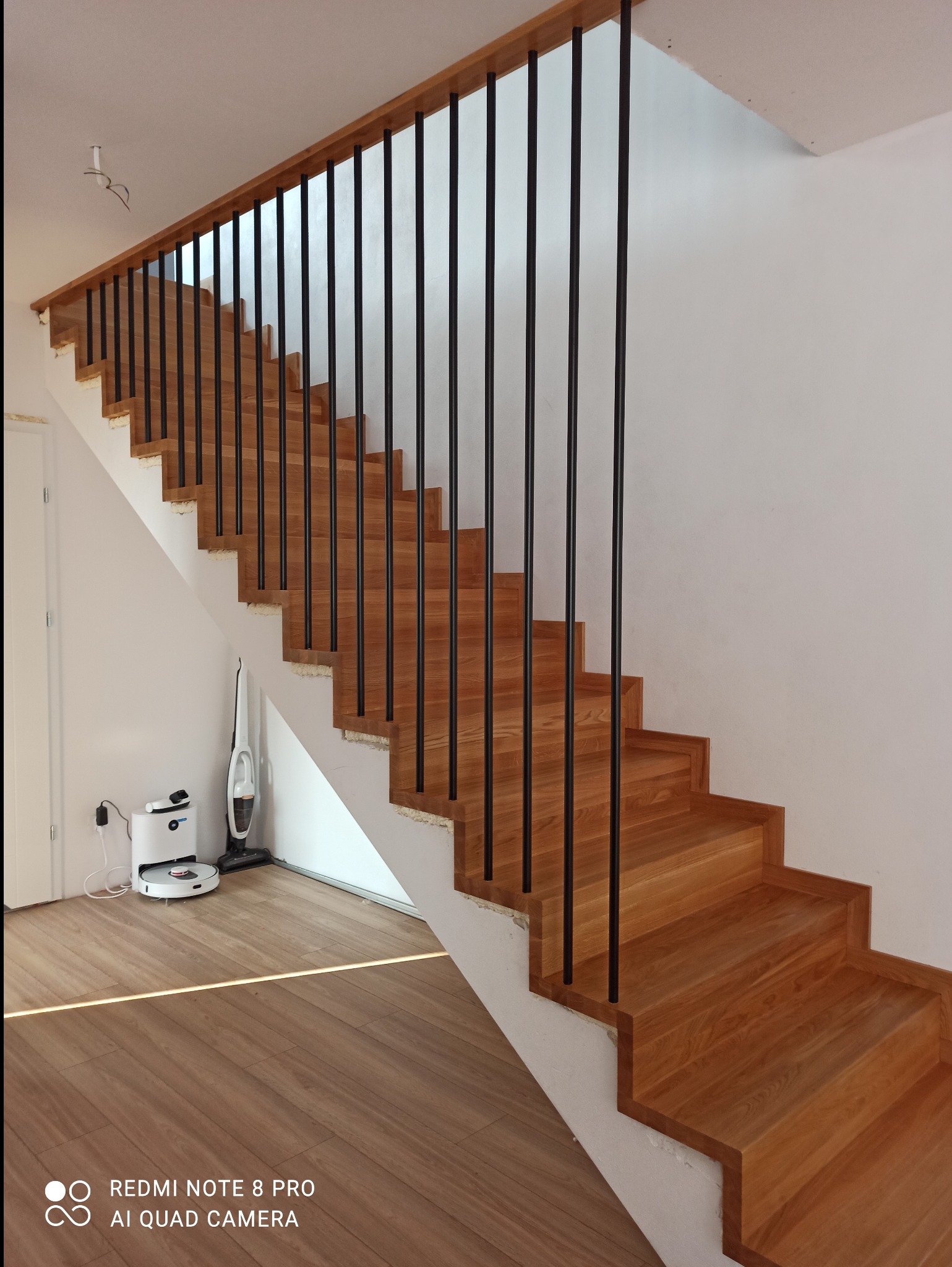 Jasne, drewniane schody z czarną, metalową balustradą w nowoczesnym wnętrzu. U dołu widoczny robot sprzątający i listwa LED w podłodze.