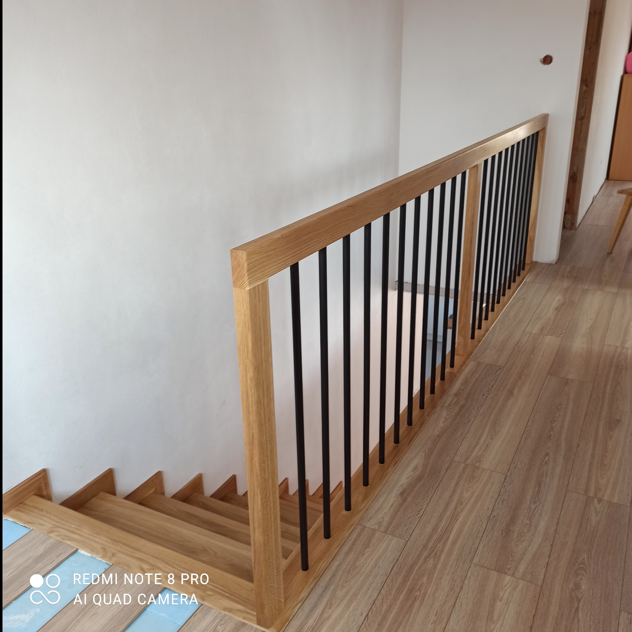 Jasne, drewniane schody z czarną, metalową balustradą, widziane z góry. Fragment podłogi wyłożonej panelami. Biała ściana w tle. Widoczne zabezpieczenie schodów.