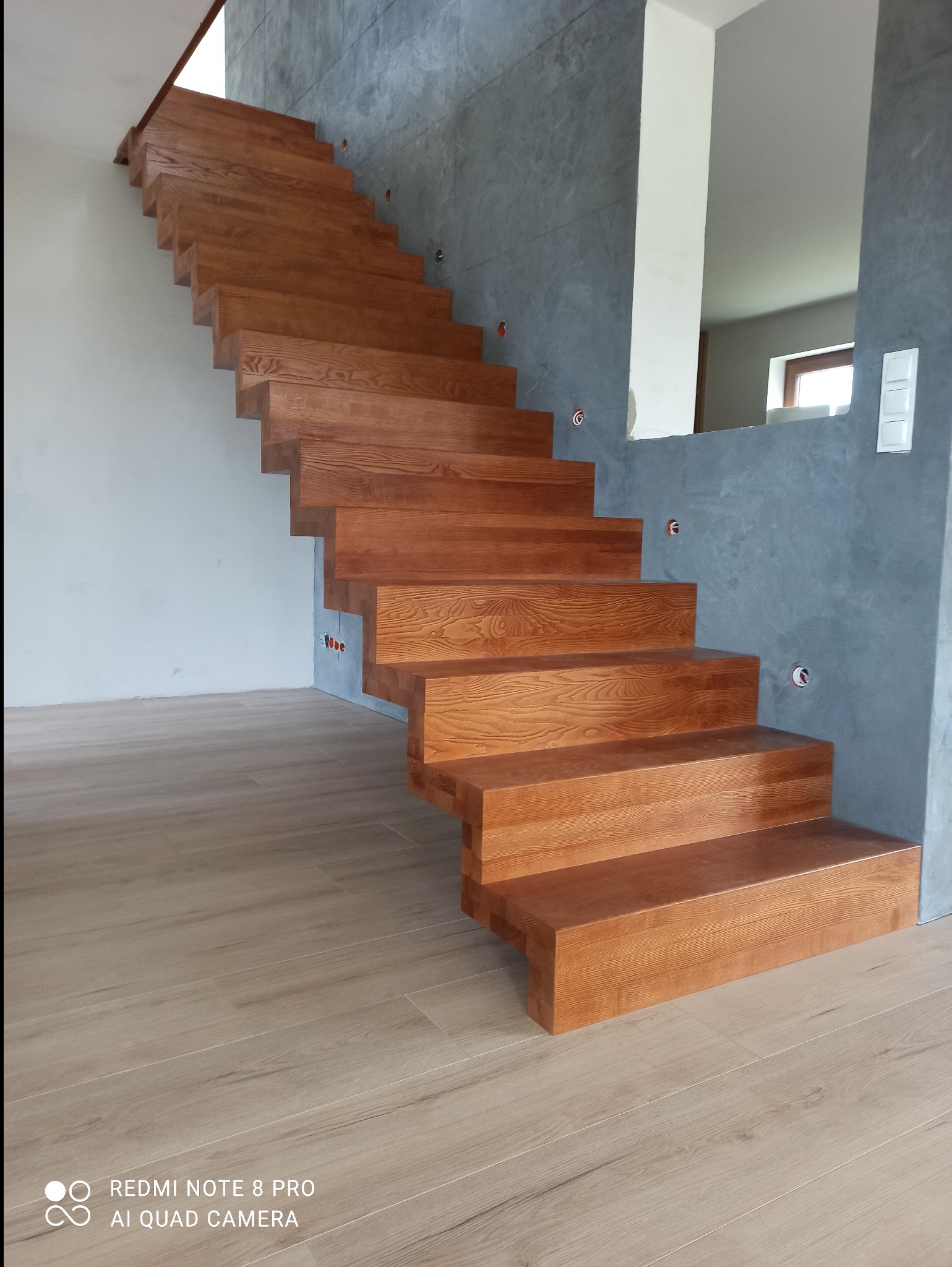 Nowoczesne, dębowe schody wspornikowe bez balustrad, z widocznym usłojeniem drewna, na tle szarej ściany z fakturą betonu i jasnej podłogi. Widoczne gniazdka elektryczne.