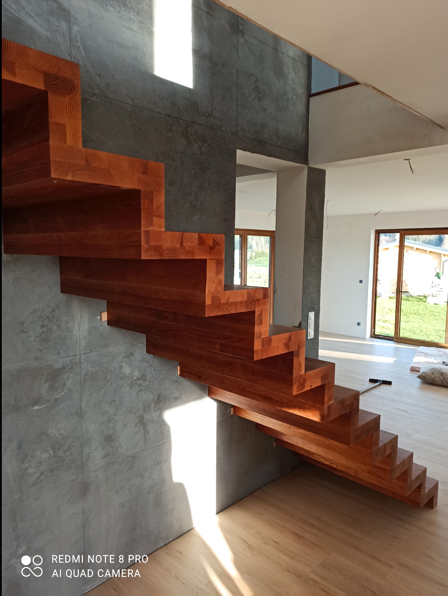Nowoczesne, drewniane schody wspornikowe z litego drewna w Naprawie, zamontowane przy szarej, betonowej ścianie. Widoczne detale usłojenia drewna i minimalistyczny design.