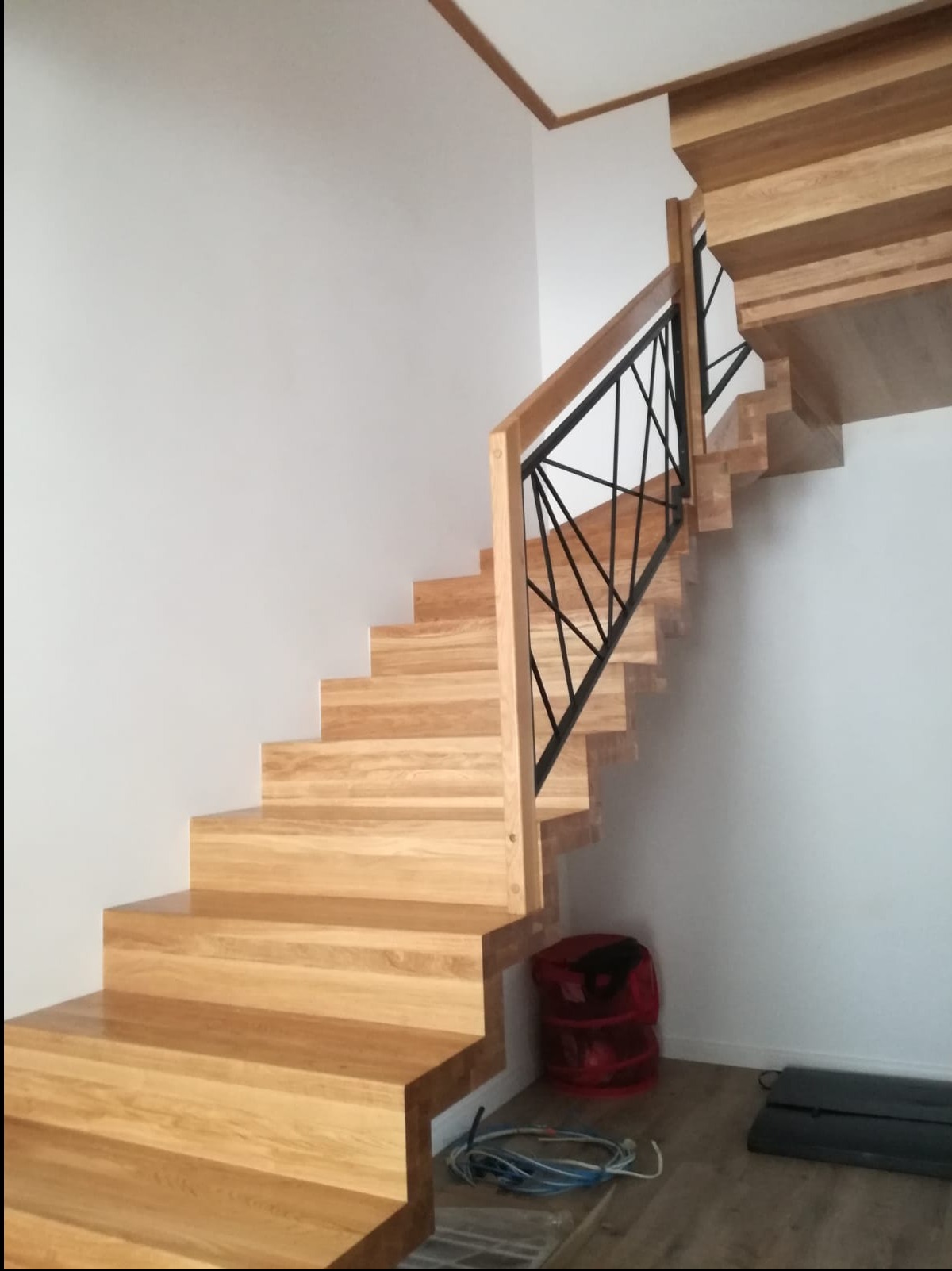 Jasne schody z drewna z czarną, geometryczną balustradą. Widoczne przewody i czerwony worek pod schodami. Minimalistyczne wnętrze z drewnianym wykończeniem sufitu.